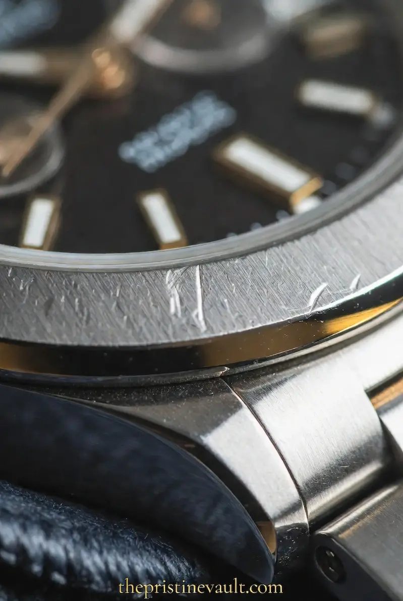 Macro close-up of visible scratches and scuff marks on a stainless steel watch bezel with a black dial, resting on a dark microfiber cloth