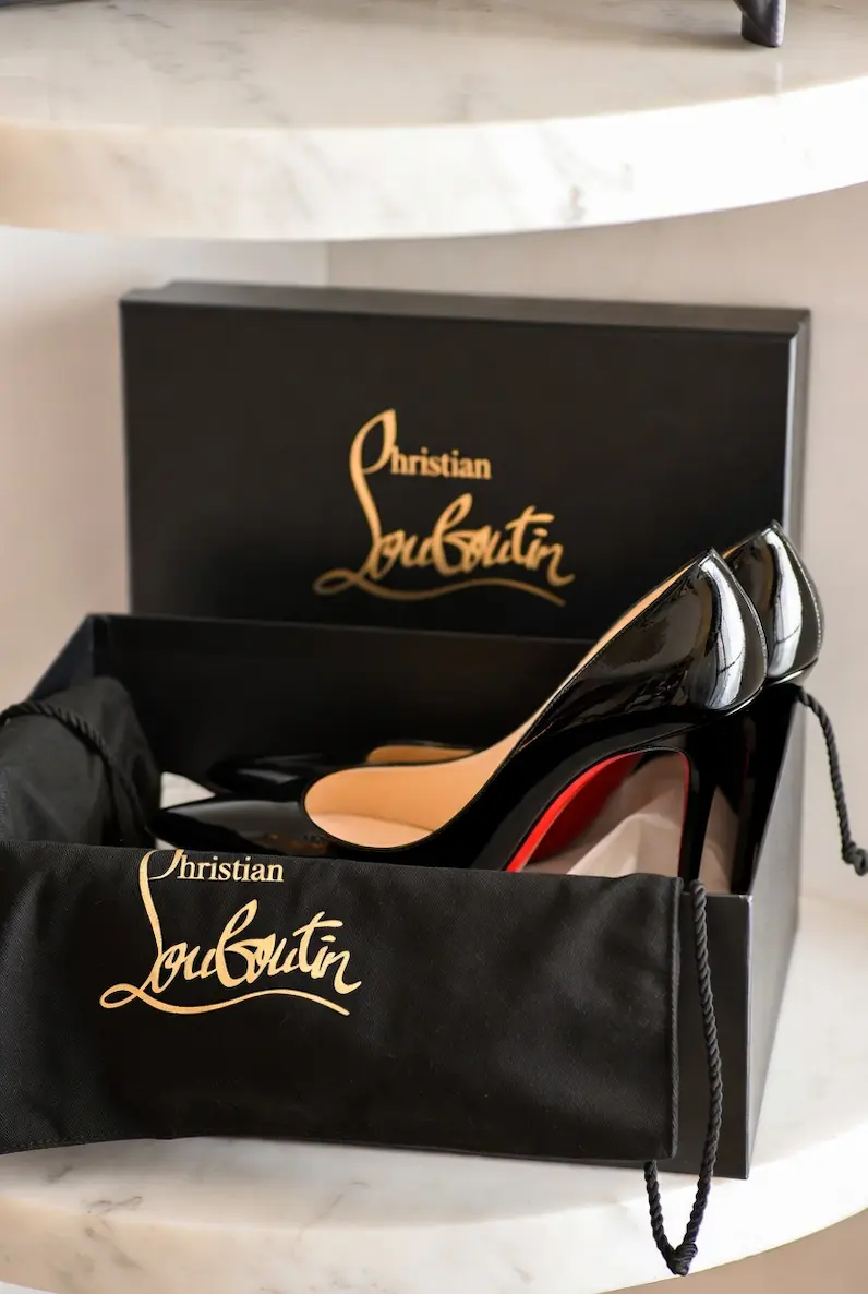 A pair of Christian Louboutin black patent leather pumps with iconic red soles resting inside the original black box and branded dust bag on a marble surface