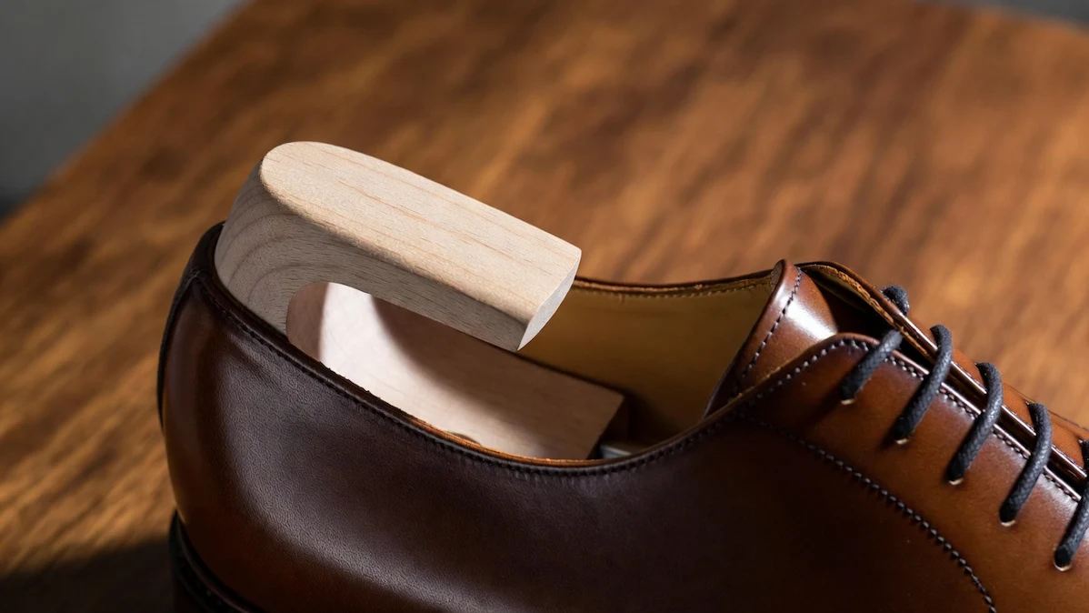 A wooden shoe tree inserted into a tan leather Allen Edmonds oxford dress shoe, resting on a warm mahogany wooden surface