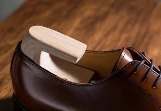 A wooden shoe tree inserted into a tan leather Allen Edmonds oxford dress shoe, resting on a warm mahogany wooden surface
