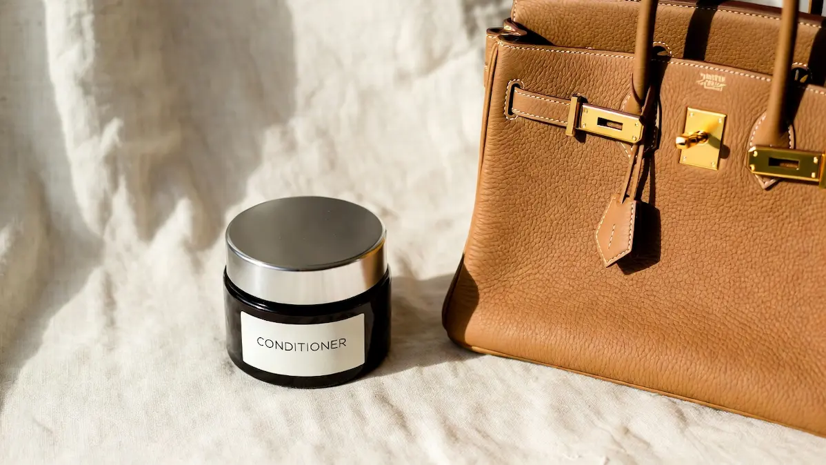 A jar of leather conditioner beside a tan Hermès Birkin bag with gold hardware on a white linen surface, natural sunlight