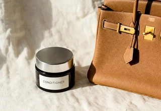 A jar of leather conditioner beside a tan Hermès Birkin bag with gold hardware on a white linen surface, natural sunlight