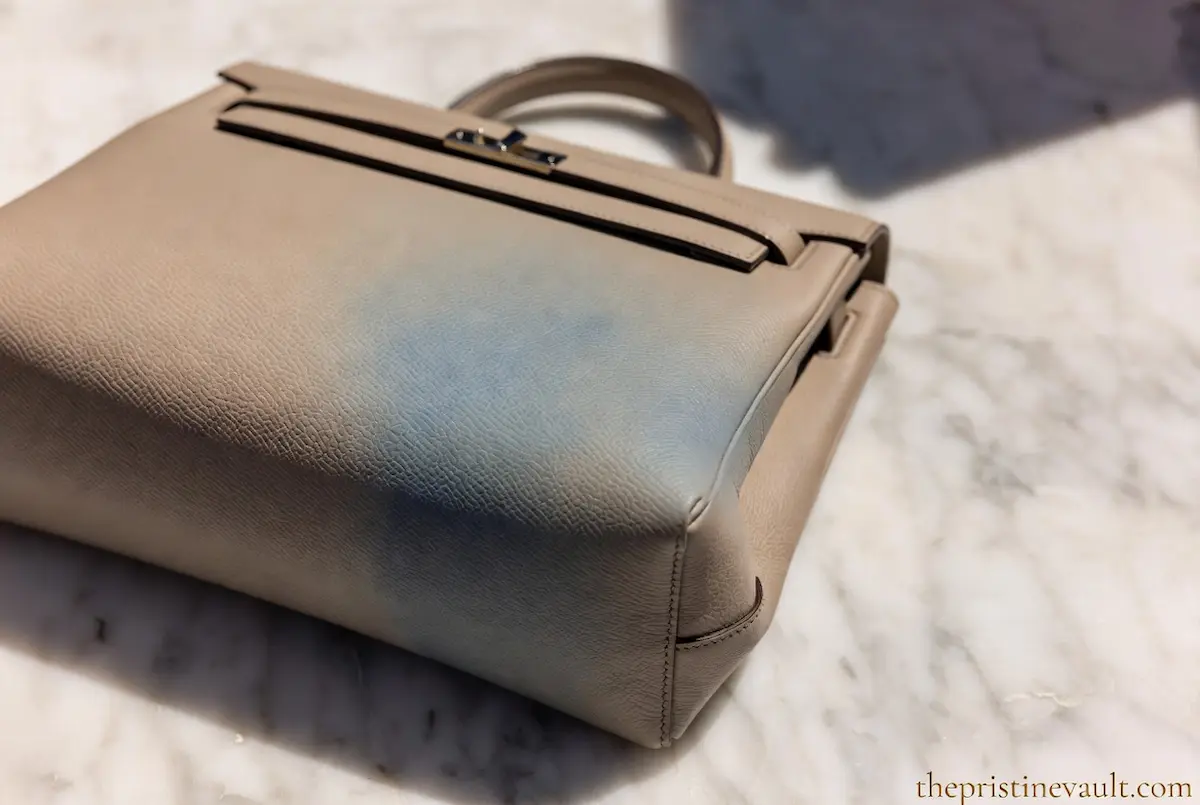 A light beige luxury leather Kelly bag with a visible blue denim color transfer stain on the side, demonstrating a leather care problem.