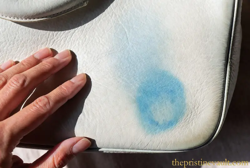 A close-up of a hand pointing to a blue denim color transfer stain on a white pebbled leather handbag, showing the texture of the damage.