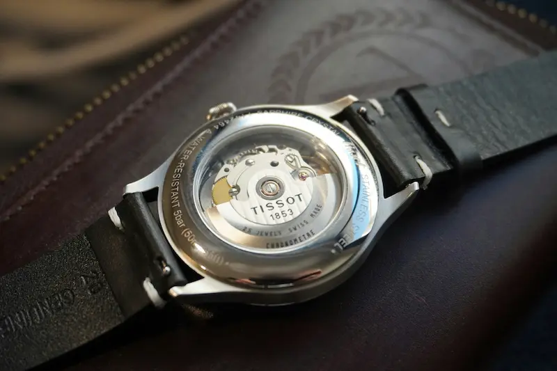 Tissot automatic watch showing open caseback with visible mechanical movement on leather surface