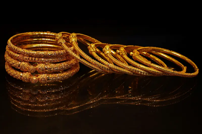 Collection of ornate gold bangles with intricate engraved patterns displayed on a reflective black surface, representing fine gold jewelry that benefits from regular tarnish removal and proper home cleaning care