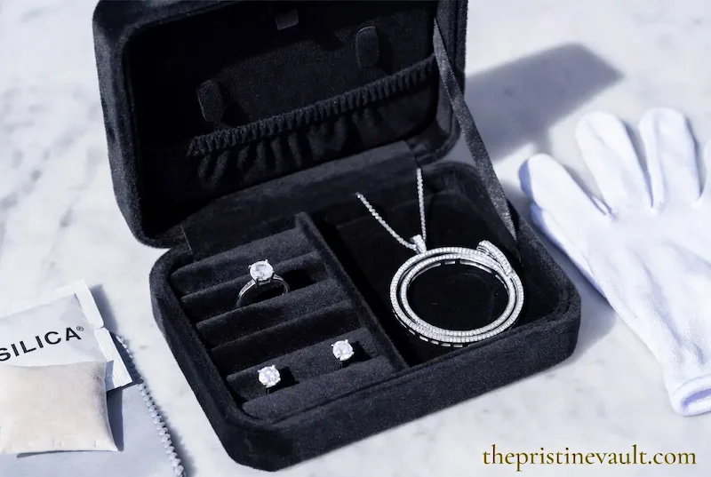 An open black velvet travel jewelry box on a marble surface displaying a diamond solitaire ring, diamond stud earrings, a diamond pendant necklace, and a diamond bangle, beside a silica gel packet and white cotton jewelry handling gloves