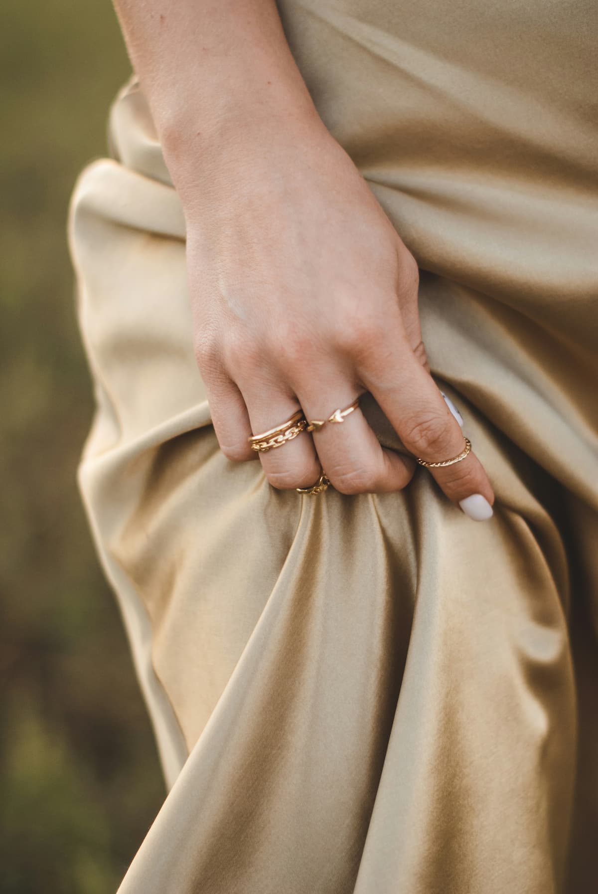 A woman's hand wearing multiple gold rings while holding a beige satin dress, illustrating elegant jewelry styling