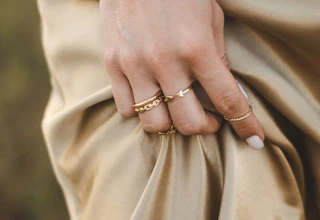 A woman's hand wearing multiple gold rings while holding a beige satin dress, illustrating elegant jewelry styling