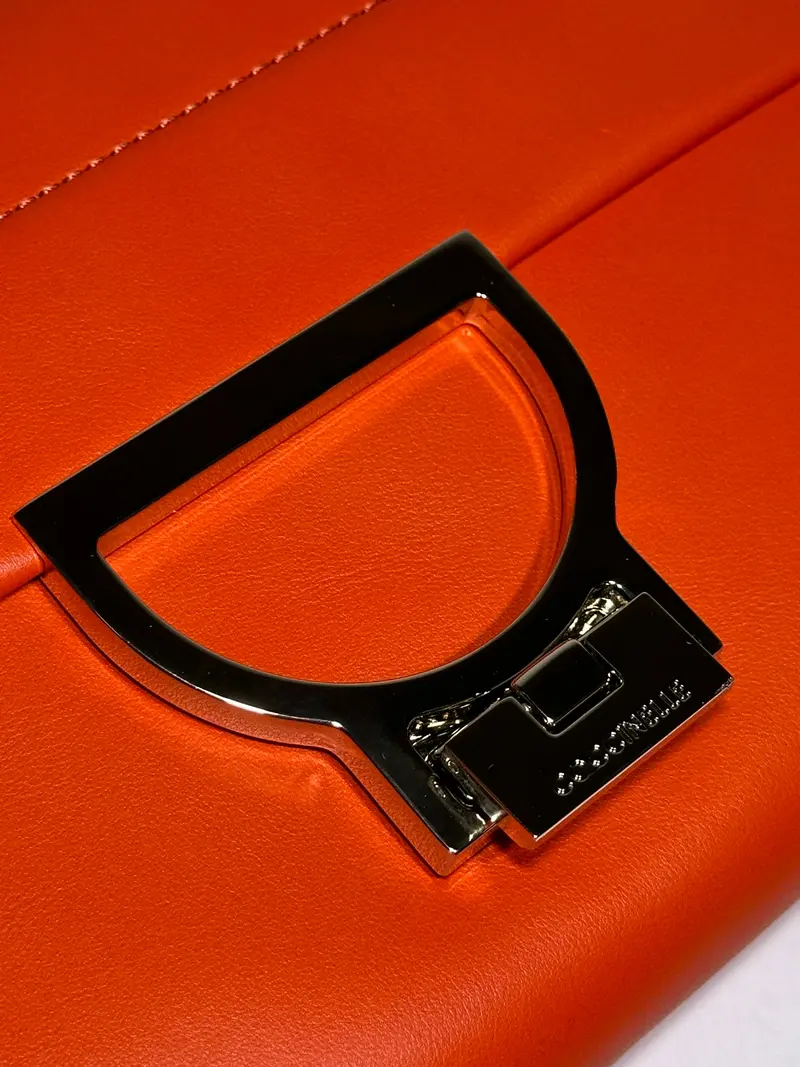 Close-up of a silver D-shaped metal clasp on a vibrant orange leather luxury handbag, showcasing premium craftsmanship