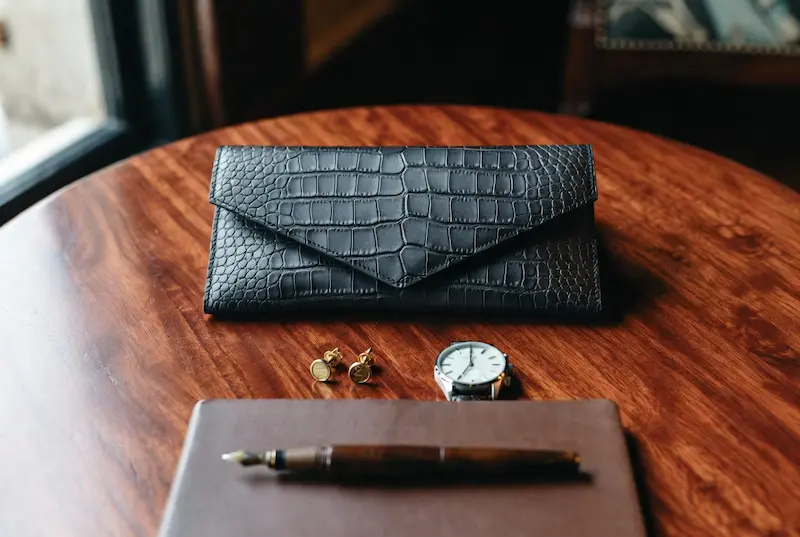 A curated lifestyle flat lay of a minimalist black crocodile leather clutch on a mahogany table next to a luxury watch and gold accessories, representing the aesthetic of quiet luxury brands like The Row or Bottega Veneta.