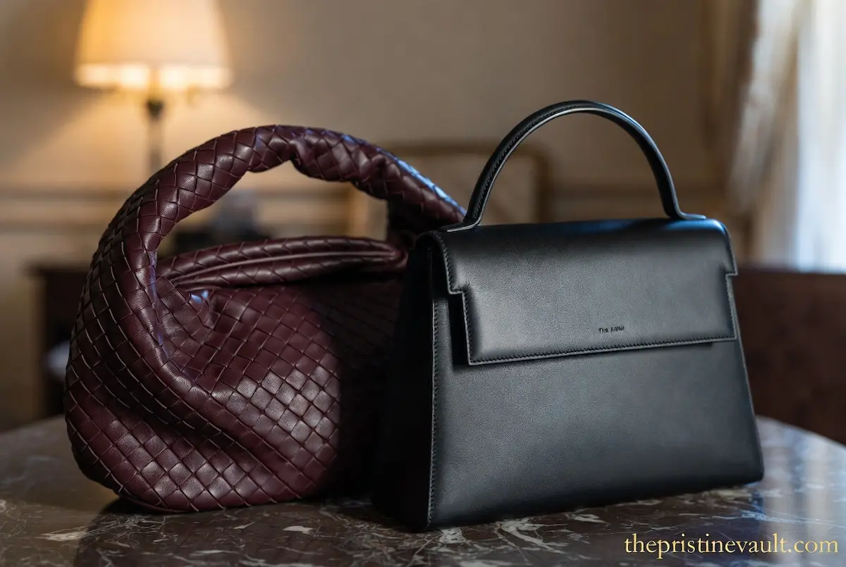 Side-by-side comparison of a deep burgundy leather woven Bottega Veneta handbag and a structured black minimalist The Row tote bag on a marble table.