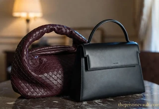 Side-by-side comparison of a deep burgundy leather woven Bottega Veneta handbag and a structured black minimalist The Row tote bag on a marble table.