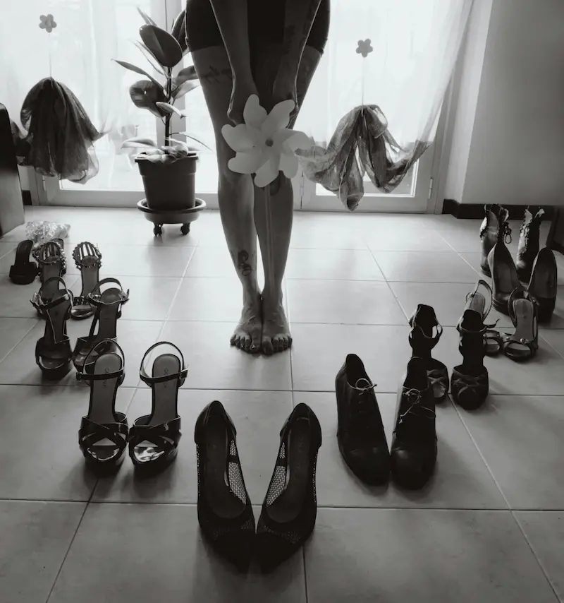 A black and white photo of a woman standing barefoot surrounded by multiple pairs of elegant heels and sandals on a tiled floor, choosing her perfect luxury shoes