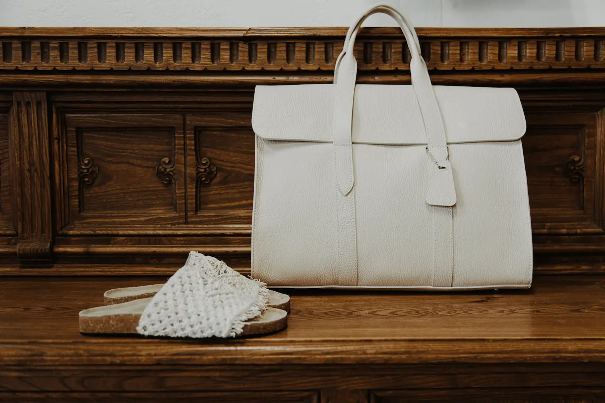 A cream leather designer tote bag and woven sandals placed on a vintage wooden dresser, illustrating proper long-term designer bag storage