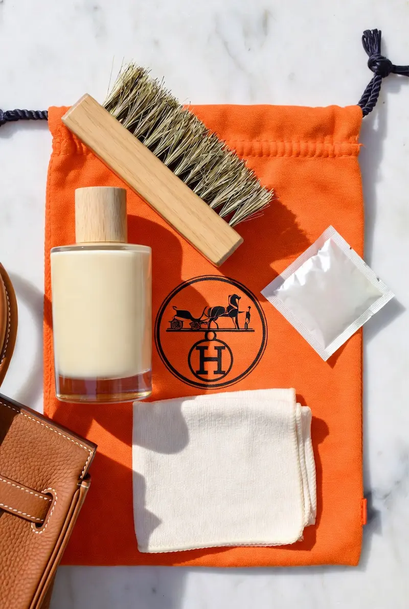 Flat lay of Hermes bag care essentials arranged on an iconic orange Hermes dust bag — leather conditioner bottle, horsehair brush, silica gel packet, and white microfiber cloth, with a tan Togo leather Birkin visible on the side