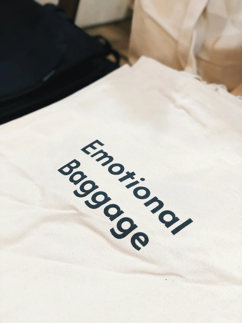 A cream canvas dust bag with 'Emotional Baggage' text, representing the type of protective dust bag used for proper Hermès bag storage and care