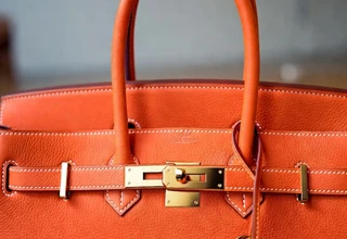 Close-up of a Hermes Birkin bag in iconic orange Togo leather with gold hardware, featuring the signature turn-lock closure, white contrast stitching, and leather clochette with gold key