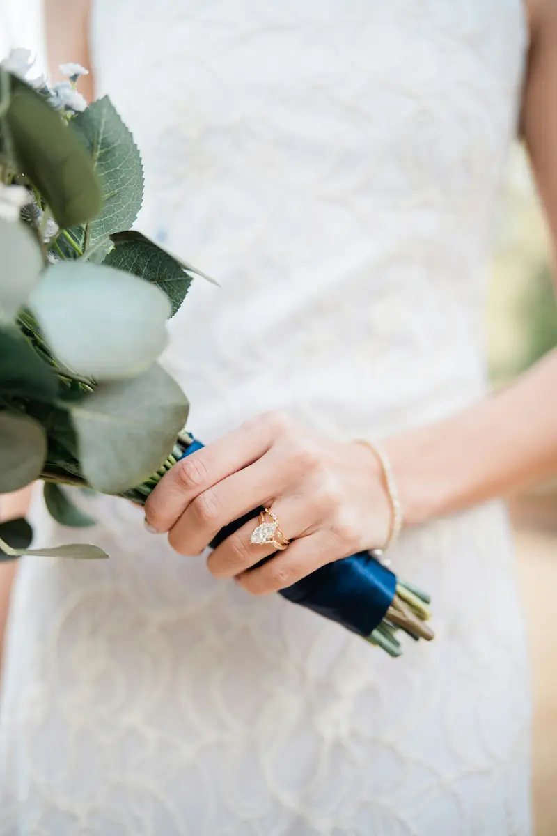 A bride wearing a pear-shaped diamond ring on a gold band while holding a green eucalyptus bouquet with navy ribbon, illustrating the importance of fine jewelry care during special occasions