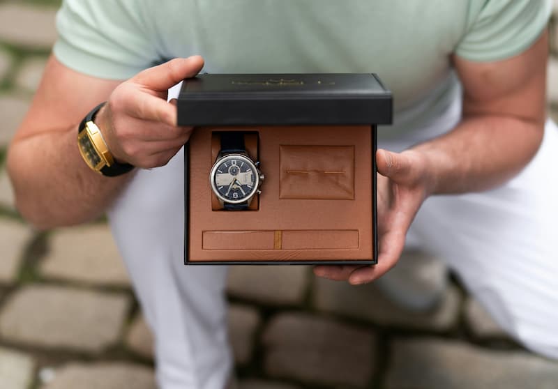 A man holding open a black luxury watch box with tan leather interior displaying a chronograph watch, showcasing premium watch storage packaging