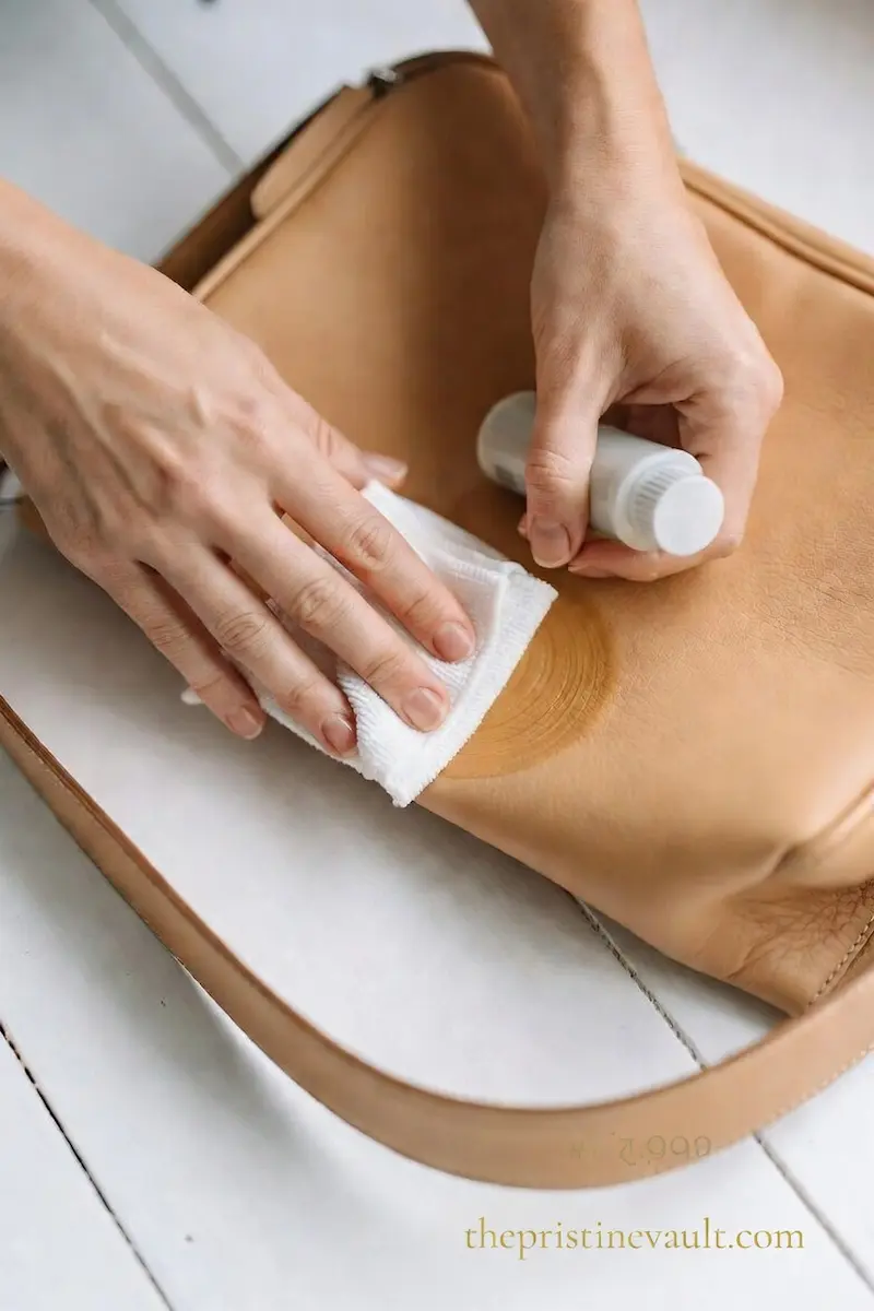 Hands applying leather conditioner with a white cloth onto a Dooney & Bourke tan vachetta leather bag on a white wooden surface