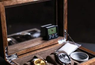 A DIY watch humidity box made from walnut wood with a glass lid, housing two luxury watches on suede cushions alongside a hygrometer, silica gel packet, and small water bowl to control humidity levels