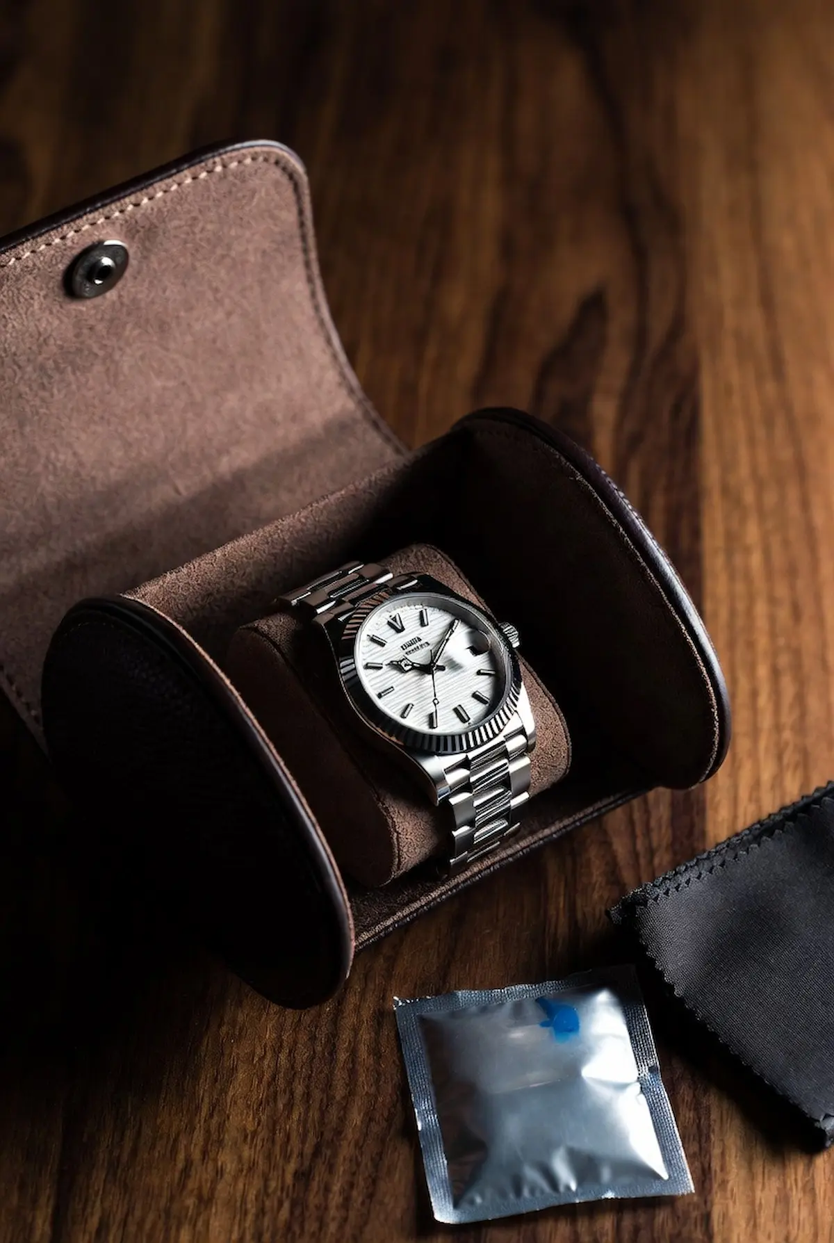 A stainless steel automatic watch with white dial stored inside a brown suede-lined leather watch roll, placed on a dark walnut surface beside a silica gel packet and black microfiber cloth