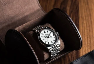 A stainless steel automatic watch with white dial stored inside a brown suede-lined leather watch roll, placed on a dark walnut surface beside a silica gel packet and black microfiber cloth