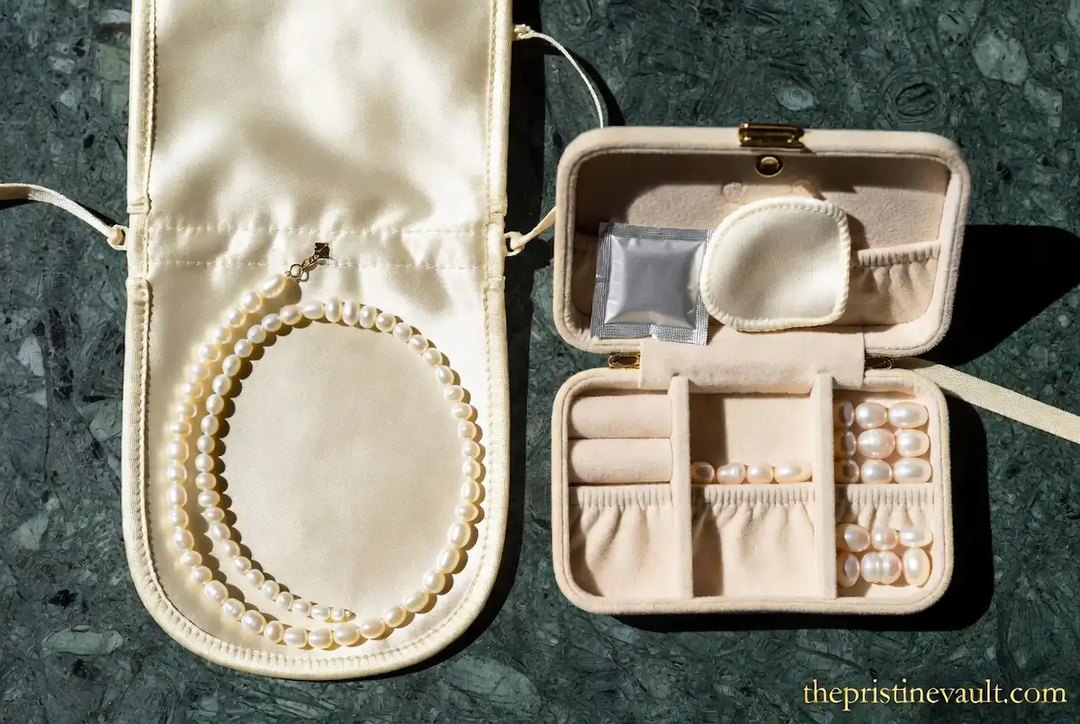A white pearl necklace coiled inside an open cream satin jewelry pouch beside a velvet travel jewelry box containing loose pearls, pearl rings, a silica gel packet, and a soft polishing pad, displayed on a green marble surface in natural sunlight