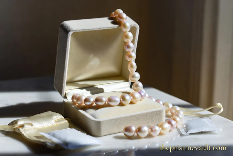 A pink freshwater pearl necklace draped over an open cream velvet jewelry box on a marble surface, beside a silica gel packet and a cream satin jewelry pouch, lit by dramatic natural side window light