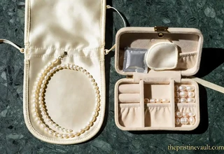 A white pearl necklace coiled inside an open cream satin jewelry pouch beside a velvet travel jewelry box containing loose pearls, pearl rings, a silica gel packet, and a soft polishing pad, displayed on a green marble surface in natural sunlight