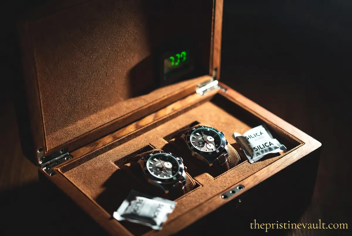 An open walnut leather watch box storing two chronograph watches on suede cushions, with silica gel packets placed in the corners and a green digital hygrometer display visible in the lid, under dramatic warm side lighting