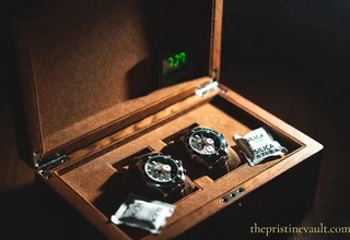 An open walnut leather watch box storing two chronograph watches on suede cushions, with silica gel packets placed in the corners and a green digital hygrometer display visible in the lid, under dramatic warm side lighting