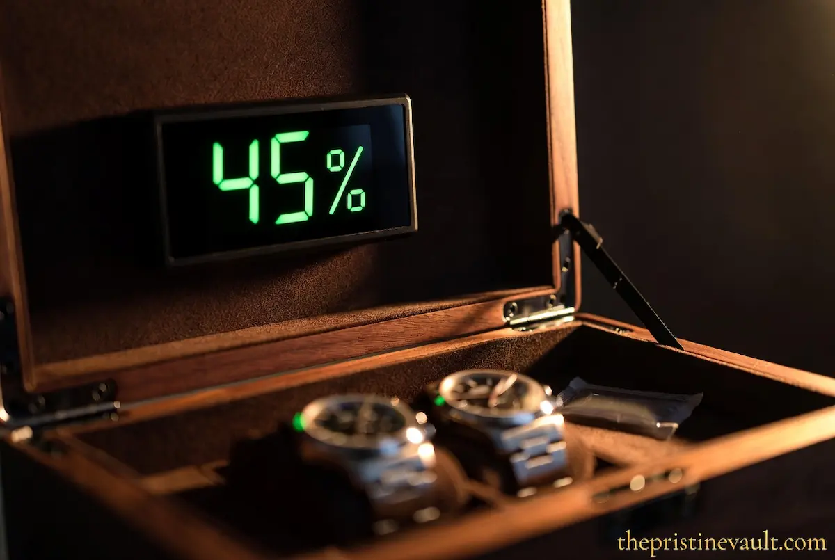 A digital hygrometer mounted inside the lid of a walnut watch storage box displaying an optimal 45% humidity reading in bright green digits, with two stainless steel watches resting on dark suede cushions below under dramatic warm side lighting