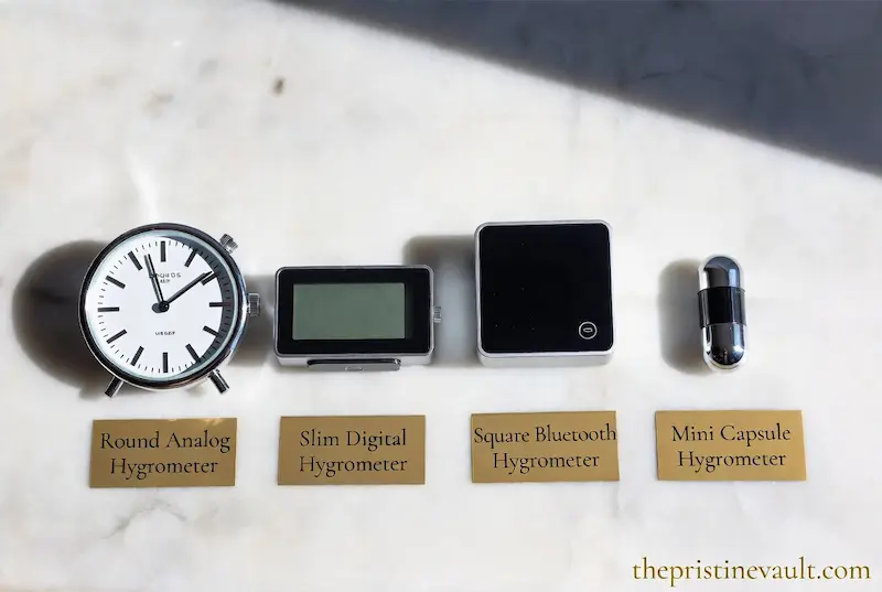 Flat lay comparison of four hygrometer types for watch storage on a white marble surface — a round analog hygrometer, a slim digital hygrometer, a square Bluetooth hygrometer, and a mini capsule hygrometer — each labeled with gold name cards in soft natural lighting