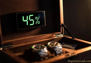 A digital hygrometer mounted inside the lid of a walnut watch storage box displaying an optimal 45% humidity reading in bright green digits, with two stainless steel watches resting on dark suede cushions below under dramatic warm side lighting