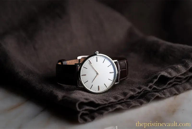 A slim minimalist quiet luxury dress watch with clean white dial and dark brown crocodile leather strap resting on folded dark linen cloth on marble surface, embodying understated elegance and stealth wealth aesthetic for men