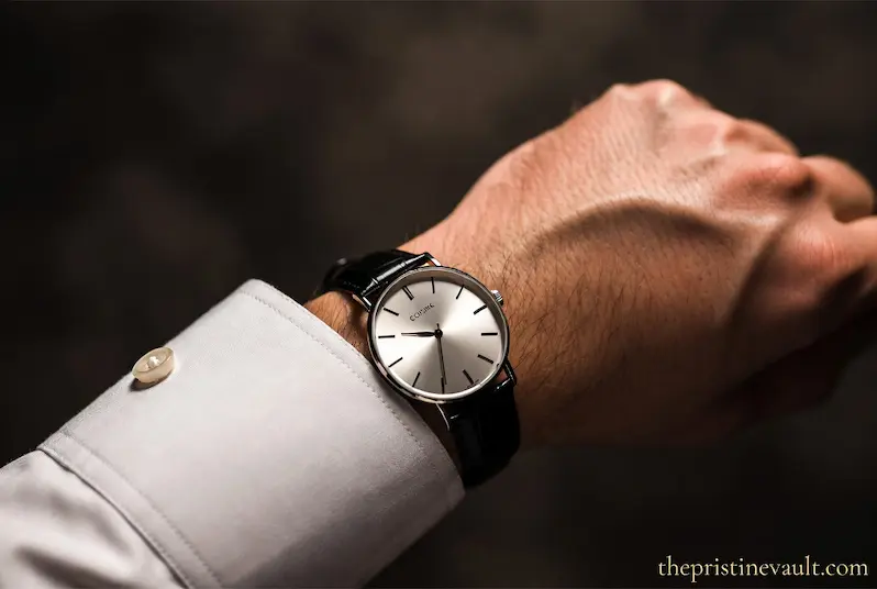 Gentleman wearing a slim minimalist quiet luxury dress watch with silver sunburst dial and black leather strap on white dress shirt cuff, representing understated stealth wealth aesthetic for men