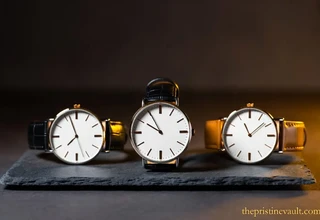 Three minimalist quiet luxury dress watches with clean white dials and slim cases displayed on dark slate, featuring black and tan leather straps, representing the best understated luxury timepieces for men