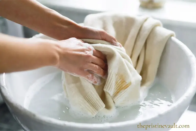 Hands gently hand washing a delicate cream cashmere sweater in white ceramic basin with mild soapy water using careful gentle motion to prevent pilling and preserve luxury knitwear fiber quality