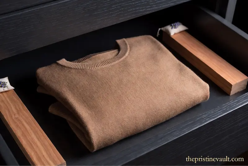 A neatly folded camel cashmere sweater stored flat inside an open dark drawer with cedar blocks and lavender sachets on both sides representing proper cashmere storage technique to prevent pilling and preserve luxury knitwear quality