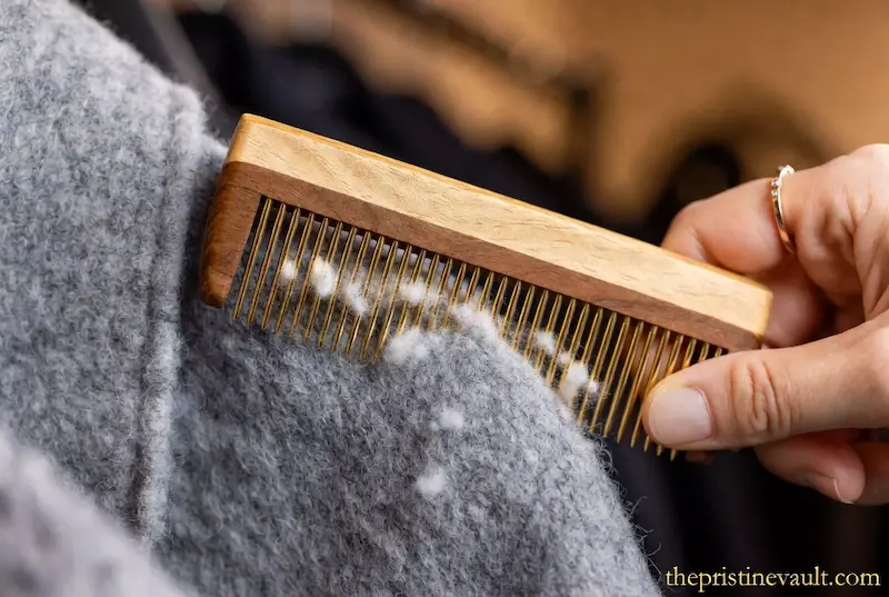 A close-up shot of a high-quality wooden cashmere comb with fine gold teeth gently removing white lint and pilling from a soft grey wool sweater.