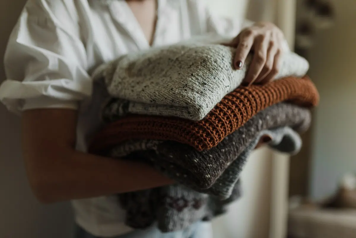 A person holding a stack of neatly folded knit cashmere sweaters in warm neutral and earth tones before putting them into moth-proof storage