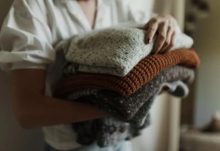 A person holding a stack of neatly folded knit cashmere sweaters in warm neutral and earth tones before putting them into moth-proof storage