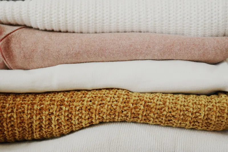 Close-up shot of a stack of clean, neatly folded cashmere and wool knit sweaters in pastel pink, white, and mustard tones