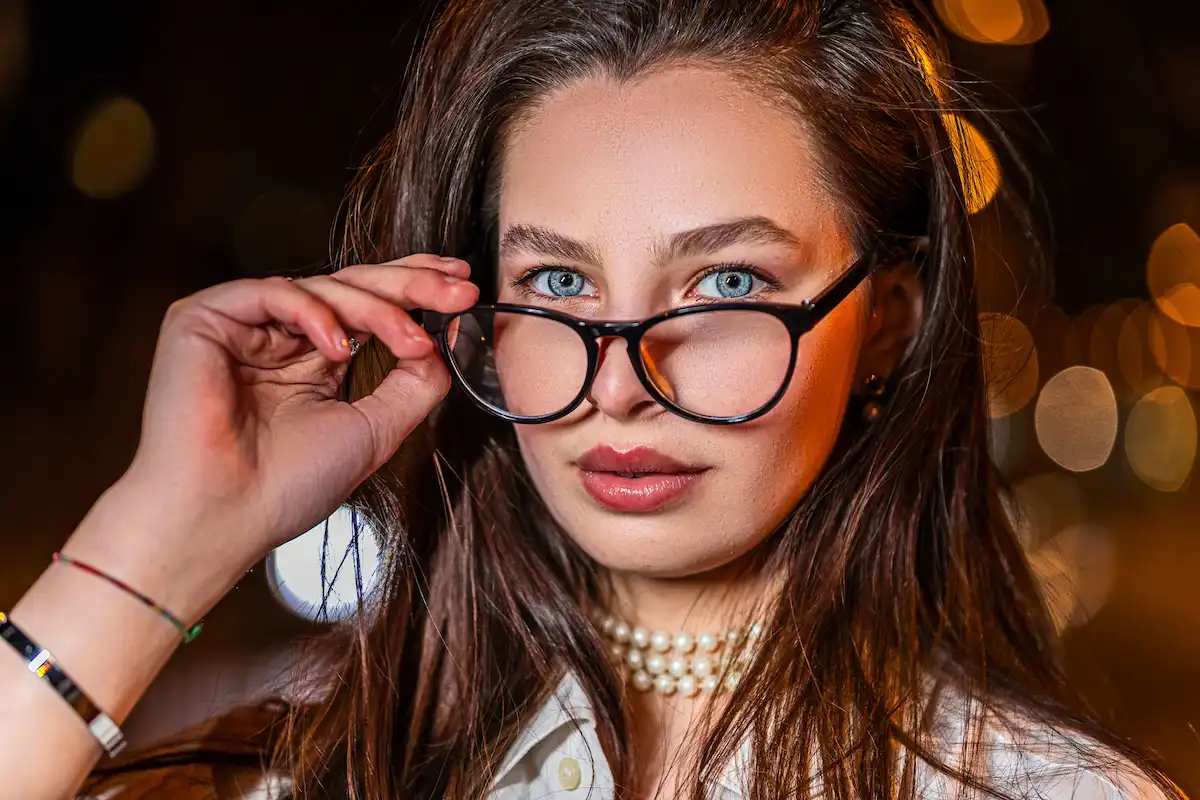 A close-up portrait of a stylish woman with blue eyes wearing elegant black luxury eyeglass frames and a pearl necklace at night