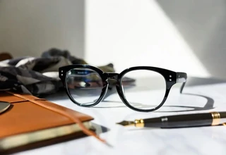 Luxury women's black acetate eyeglass frames on a white marble desk with a leather journal and fountain pen, representing high-end designer eyewear collections.