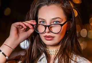 A close-up portrait of a stylish woman with blue eyes wearing elegant black luxury eyeglass frames and a pearl necklace at night