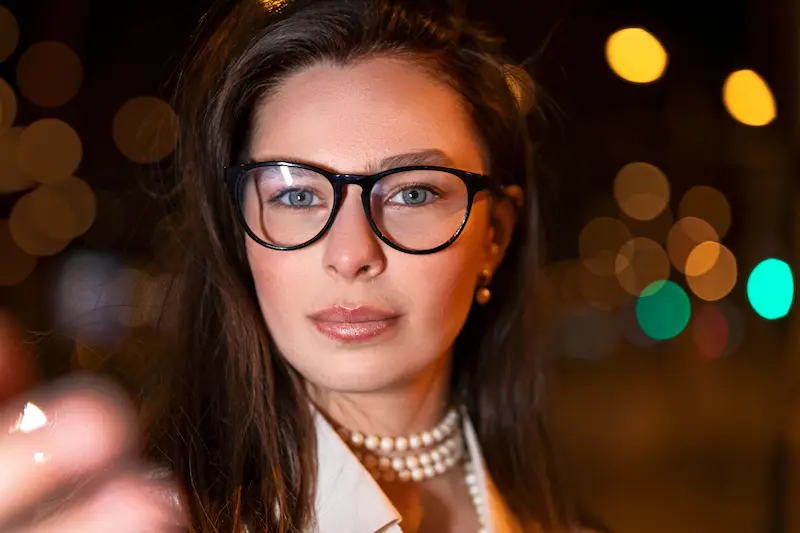 Front portrait of an attractive woman with blue eyes wearing stylish black luxury prescription eyeglasses against golden city bokeh lights