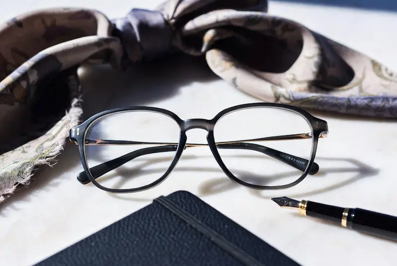 A pair of subtle black women's eyeglasses with gold details, next to a fountain pen, leather journal, and silk scarf on a white marble desk, perfect for luxury curators.